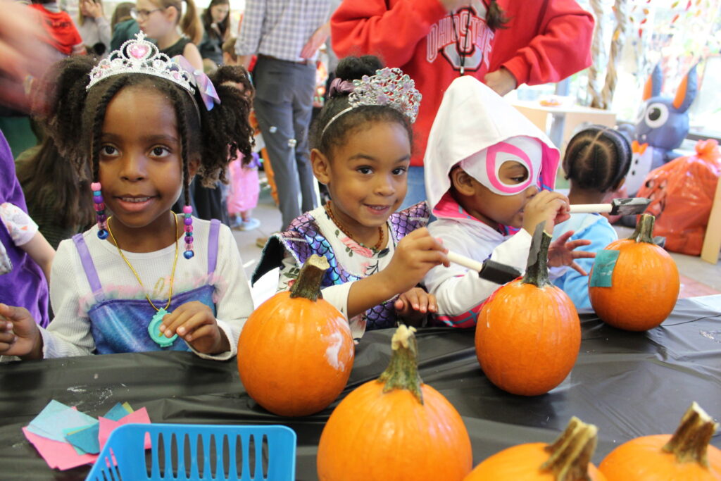 Three young children dressed as princesses stand in front of a table with pumpkins.