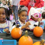 Three young children dressed as princesses stand in front of a table with pumpkins.