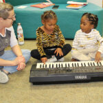 A woman is seated at left on the carpeted floor of a preschool library. She is looking at two small children, each of whom is pressing fingers onto a Yamaha electric keyboard.