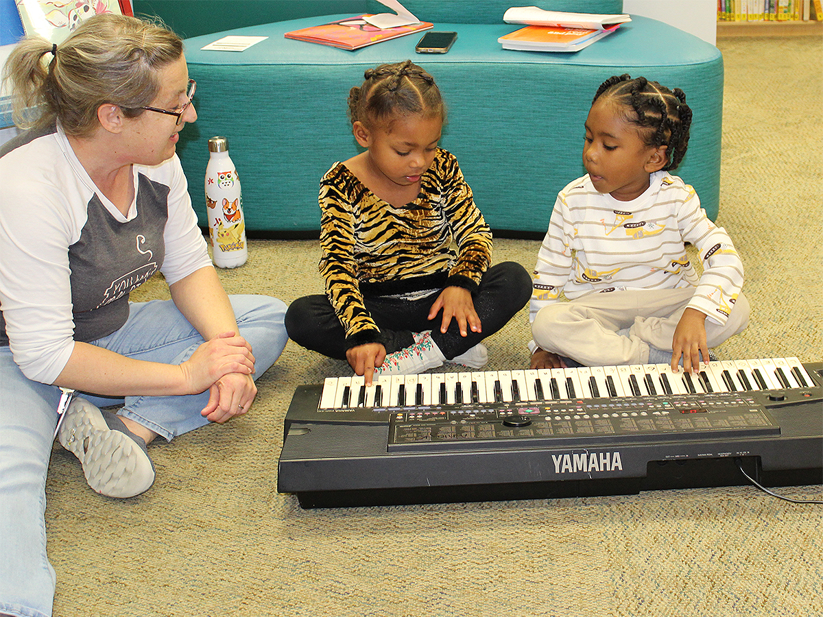 A woman is seated at left on the carpeted floor of a preschool library. She is looking at two small children, each of whom is pressing fingers onto a Yamaha electric keyboard.