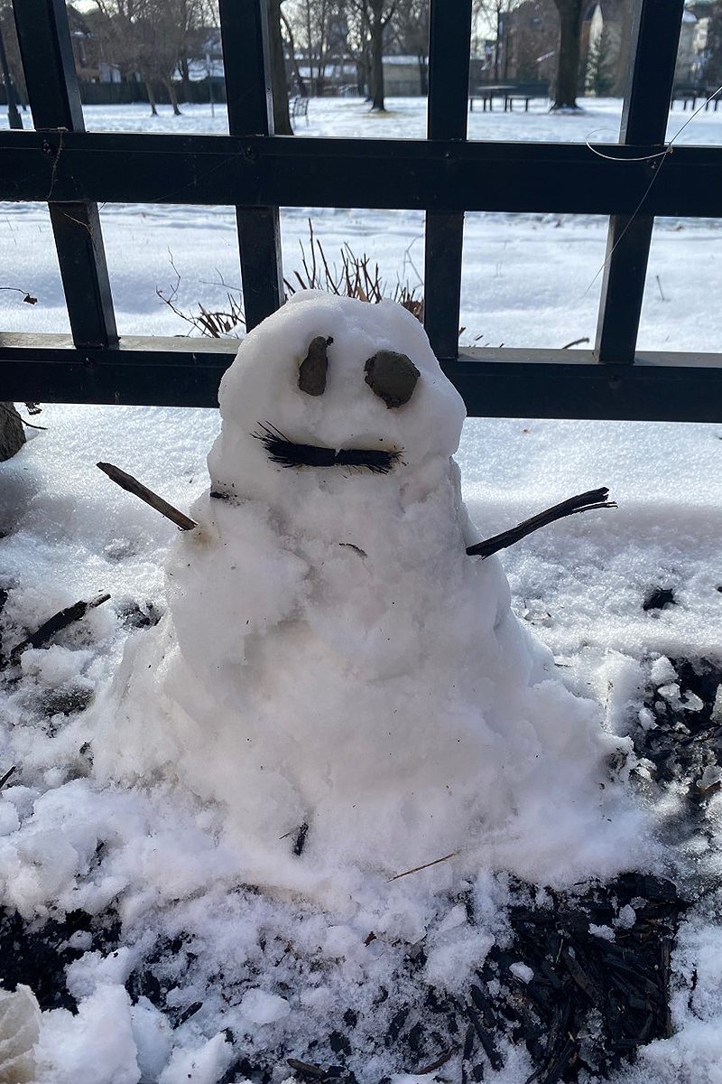 A small snowman with sticks for arms, a stick for a mouth, and two small pieces of bark for eyes, faces the camera, with a wooden fence with wide openings between the slats behind the snowman, and a snow-covered playground in the background.