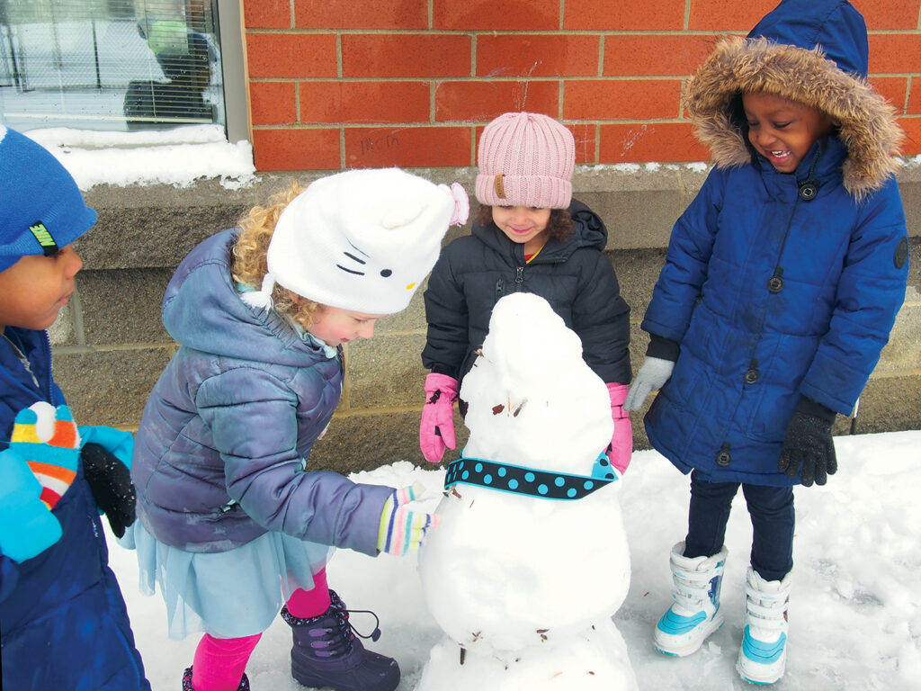 Four young children surround a snowman. The children are bundled up in winter coats, wool hats, and children's boots. The children and the snowman are next to a building made of concrete blocks and bricks. A blue and black polka dot ribbon hangs around the neck of the snowman.