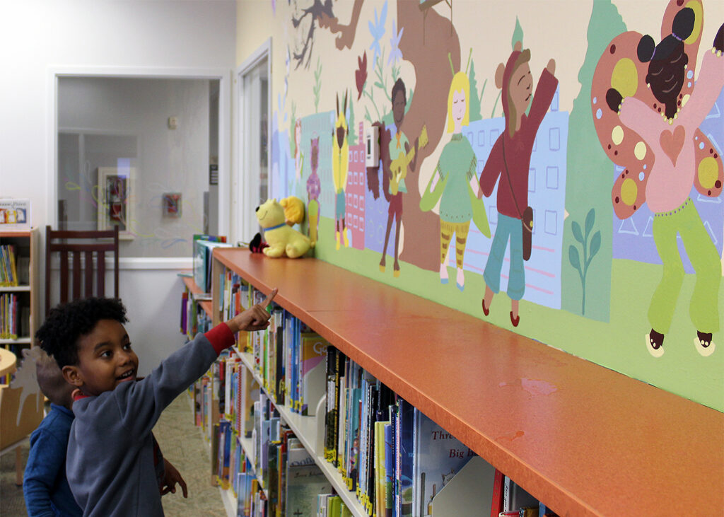 A small child points to a colorful mural on the wall of a preschool library. The colorful mural depicts smiling young children of diverse ethnicities and appearances depicted with characteristics of some animals, like butterfly wings, bear ears, rabbit ears, and insect wings and antennae.