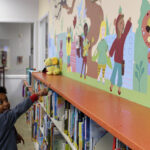 A small child points to a colorful mural on the wall of a preschool library. The colorful mural depicts smiling young children of diverse ethnicities and appearances depicted with characteristics of some animals, like butterfly wings, bear ears, rabbit ears, and insect wings and antennae.